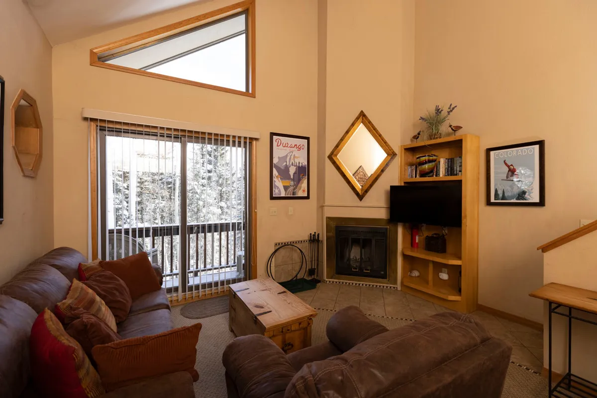 The image showcases a warm and inviting living room with a rustic fireplace topped by a wooden mantle, a comfortable leather sofa adorned with colorful pillows, and a large sliding glass door framing a snowy mountain landscape. A wooden bookshelf with a TV and tasteful decor completes the cozy mountain lodge ambiance.
