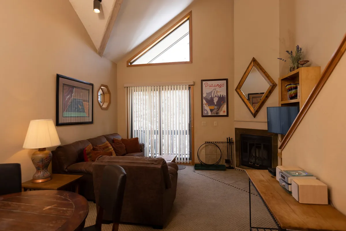 A cozy living room featuring a brown sectional sofa with plush cushions, a fireplace with fireplace tools, a decorative wall mirror with a wooden frame, and a sliding glass door with vertical blinds leading to a balcony. The space is sunlit and filled with warm, homey touches. The layout encourages relaxation, socializing, and easy access to the outdoors.