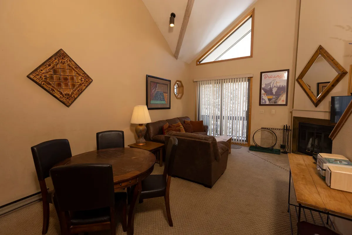 An inviting living area with a leather sectional sofa, a fireplace with wood storage, a round wooden dining table with chairs, and a large window with vertical blinds letting in abundant natural light. The space feels open and bright, ideal for both fireside relaxation and lively gatherings.