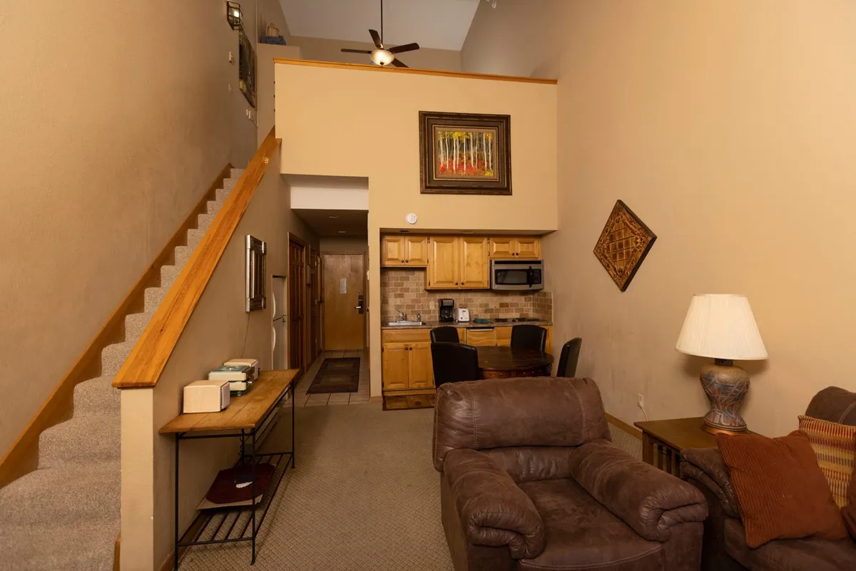 A warm, compact kitchen and living area with wooden cabinetry, decorative wall art, a plush brown armchair, and a small table, all adjacent to a carpeted staircase. The scene is bright and inviting, ideal for casual dining, morning coffee, or relaxed family time before or after a day on the slopes.