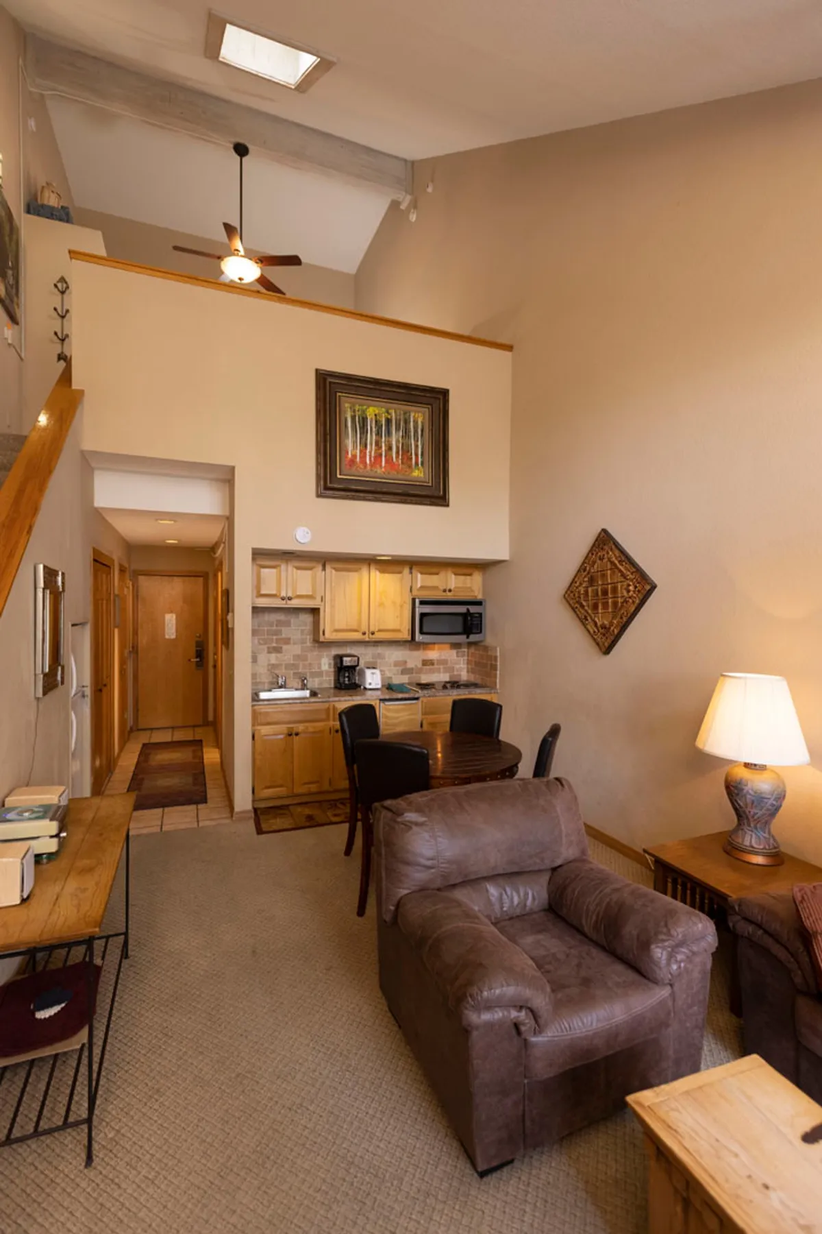 A warm and comfortable living space with a leather armchair, small wooden dining table with chairs, a kitchenette with wooden cabinets and a tiled backsplash, and artwork on the walls. The mood is inviting and homey, perfect for relaxing, dining, or playing games after a day outdoors.