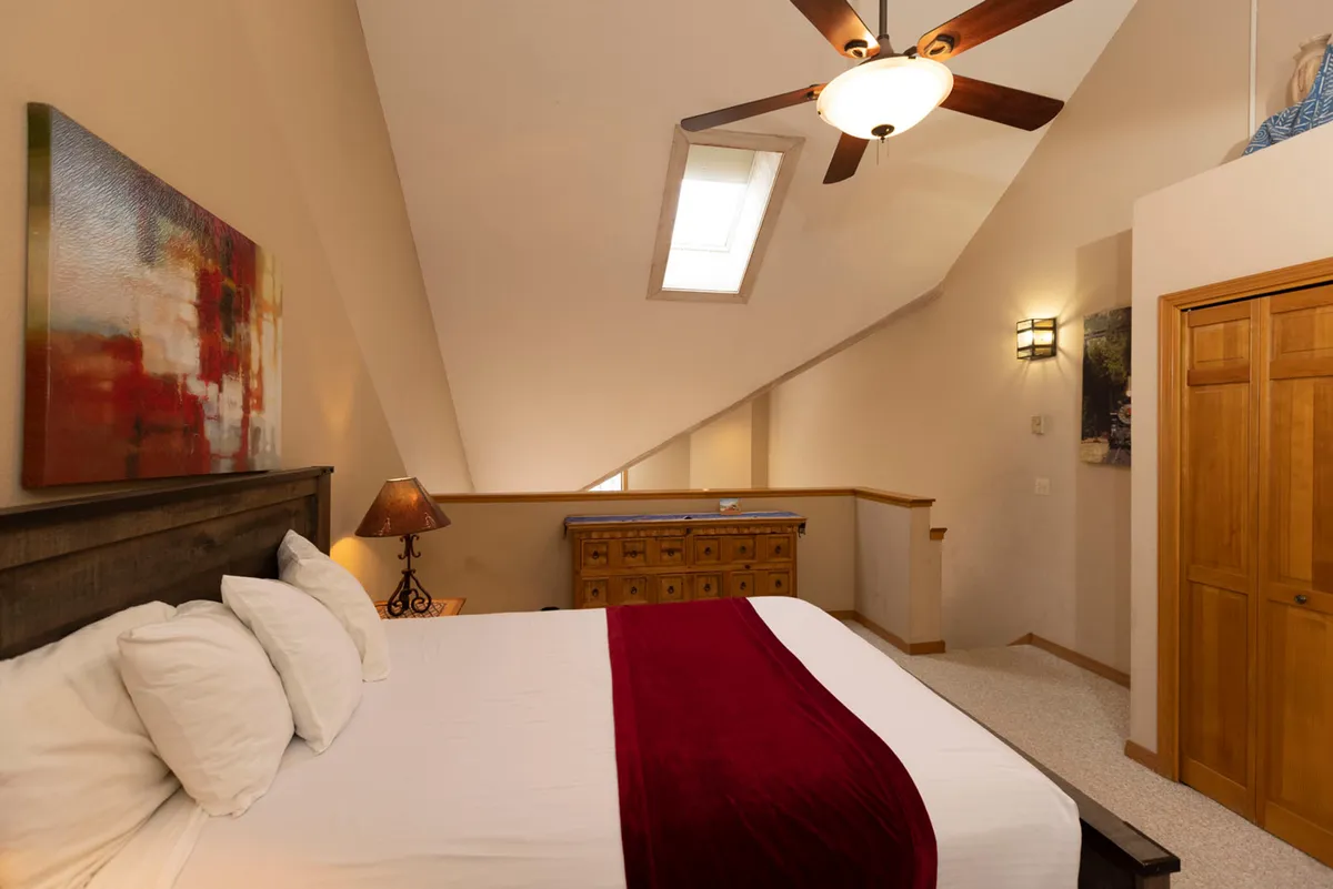 A spacious bedroom featuring a large bed with crisp white linens and a deep red runner, a wooden chest of drawers beneath a skylight that floods the room with natural light, and an abstract colorful painting hung above the bed. Wooden closet doors offer convenient storage, and the room exudes a peaceful, inviting mountain lodge ambiance.