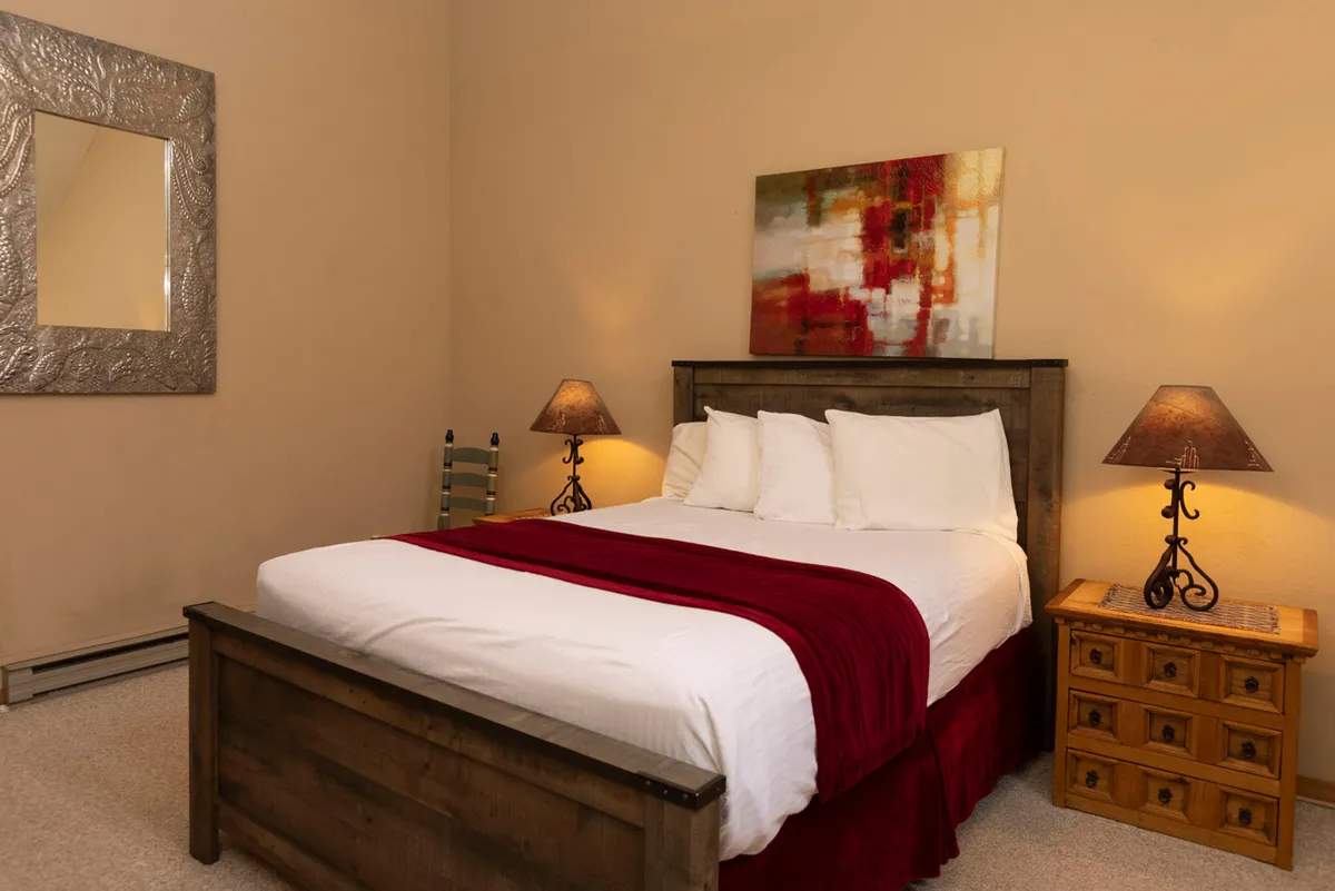 A warmly lit bedroom with a wooden bed frame dressed in white linens and a red throw, flanked by matching bedside tables with decorative lamps. An abstract painting above the bed and an ornate silver-framed mirror add touches of elegance, while the overall ambiance is inviting and restful.