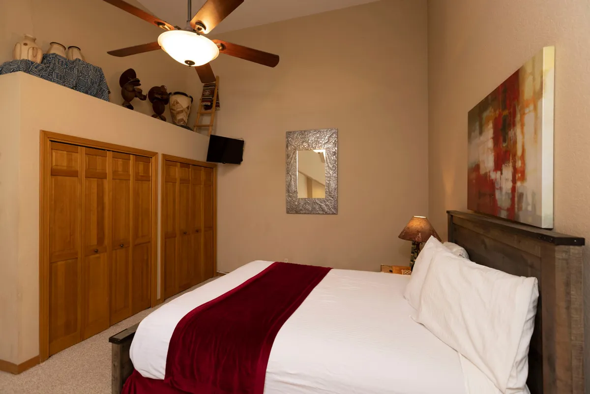 A cozy bedroom with white linens accented by a rich red throw, wooden closet doors, a decorative mirror, and an abstract painting above the bed. The setting exudes comfort and warmth, inviting guests to unwind and recharge in a private, intimate space.