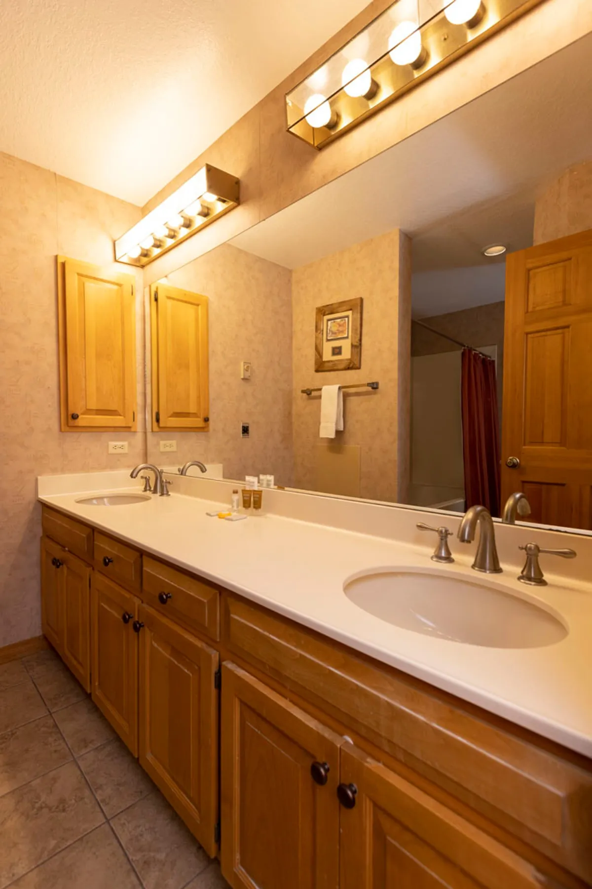 A bright, modern bathroom featuring a double sink vanity with ample countertop space, a large mirror with strong lighting, and wood cabinetry. The shower area is visible in the background, creating a sense of privacy and luxury.