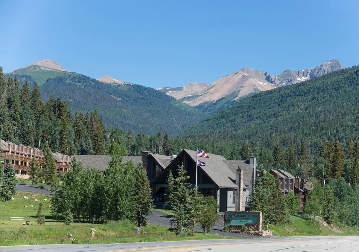 The exterior of Cascade Village lodge is featured, with its peaked roof, stone chimney, and welcoming landscaping set against a stunning mountain backdrop. The lodge sign is visible at the roadside, and the lush surroundings emphasize the resort’s connection to nature and the mountains.