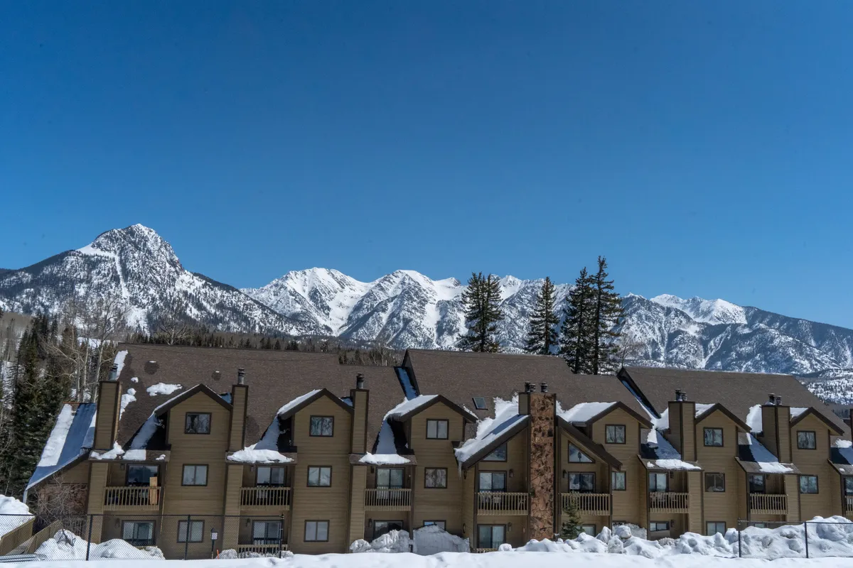 A row of chalet-style townhomes with balconies sits under snow-covered roofs, backed by a dramatic, snow-capped mountain range and clear blue sky. The scene is peaceful yet invigorating, highlighting the cozy retreat atmosphere after a day of adventure.