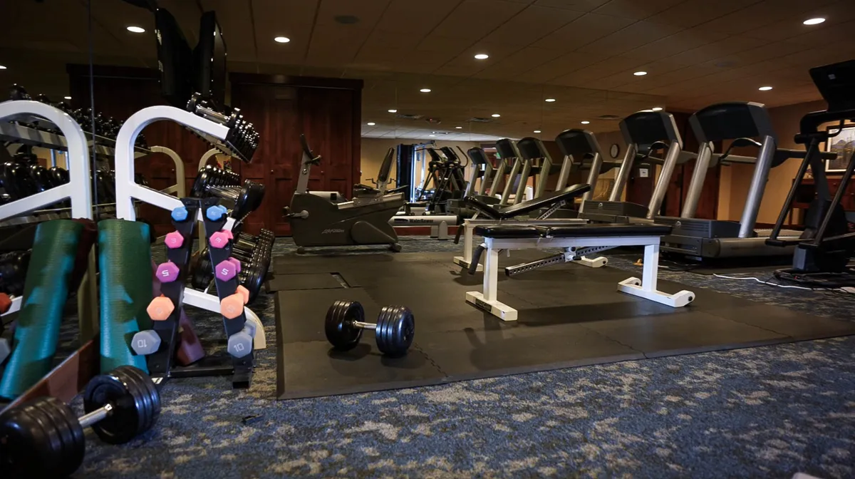 A bright, well-equipped fitness center featuring a row of treadmills, a weight bench, racks of dumbbells, and exercise mats. The modern interior suggests opportunities for guests to maintain their fitness routines while enjoying their vacation.