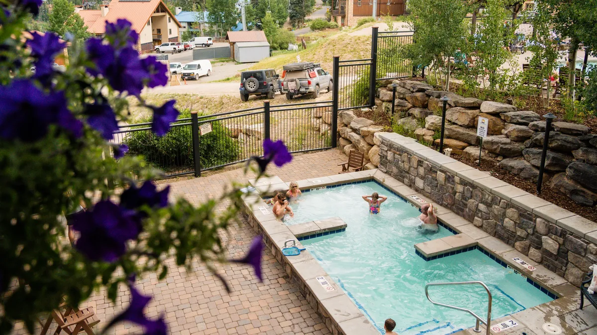 A welcoming heated outdoor pool area where guests enjoy the warm water, bordered by a stone wall and enhanced by colorful purple flowers in the foreground. The relaxed vacation vibe is complemented by nearby buildings and a sense of community.