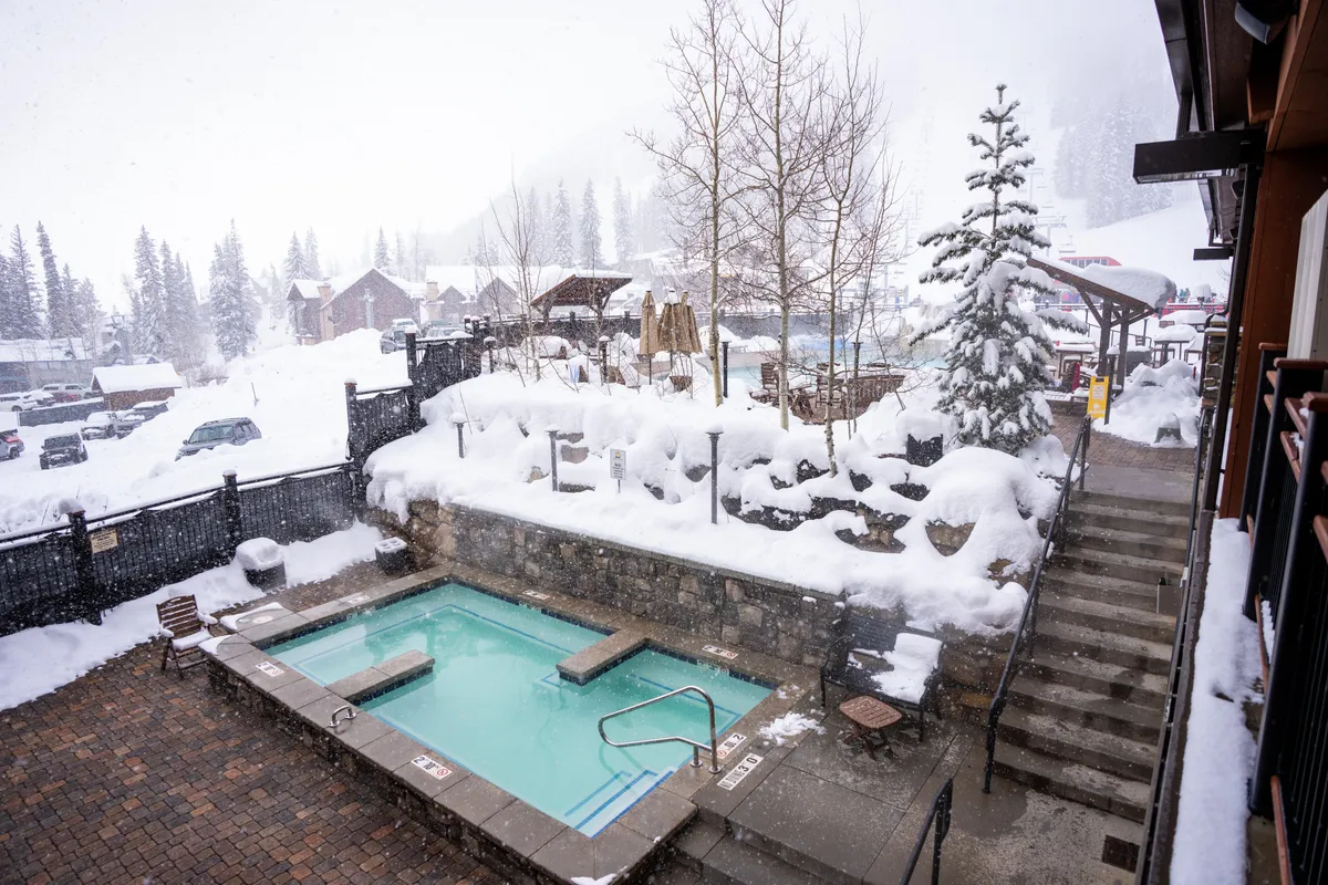 An inviting heated outdoor pool with steam rising from the water, surrounded by snow-covered stone walls and stairs. Evergreen and deciduous trees, blanketed in snow, frame the scene, while rustic wooden structures add to the cozy mountain atmosphere. The setting is tranquil, evoking both relaxation and exclusivity.