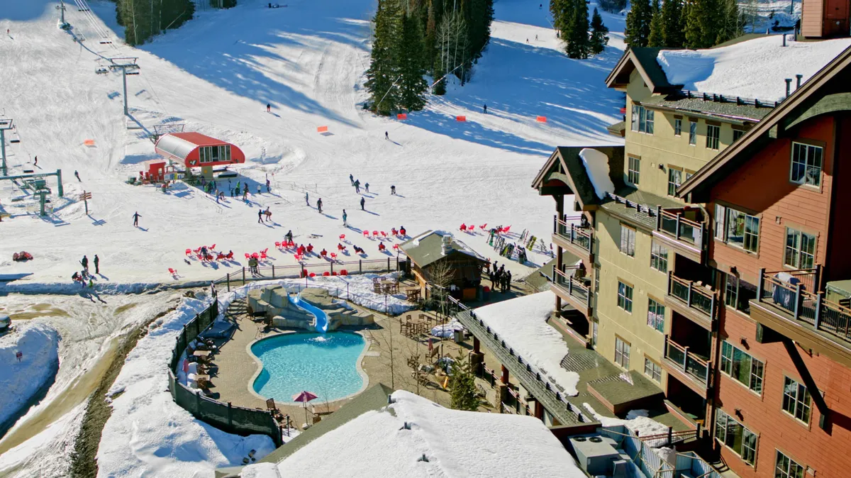 A lively scene featuring a heated outdoor pool with a water slide, set against snowy mountain slopes with visible skiers, a large multi-story lodge with balconies, and a red ski lift station. The mix of snow, water, and resort architecture highlights the year-round fun available at the property.