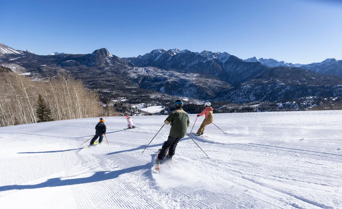 Four skiers descend a well-groomed ski slope with a snow-covered mountain range in the background and clear blue skies above. Leafless trees line the slope, underscoring the winter setting and the excitement of outdoor recreation.